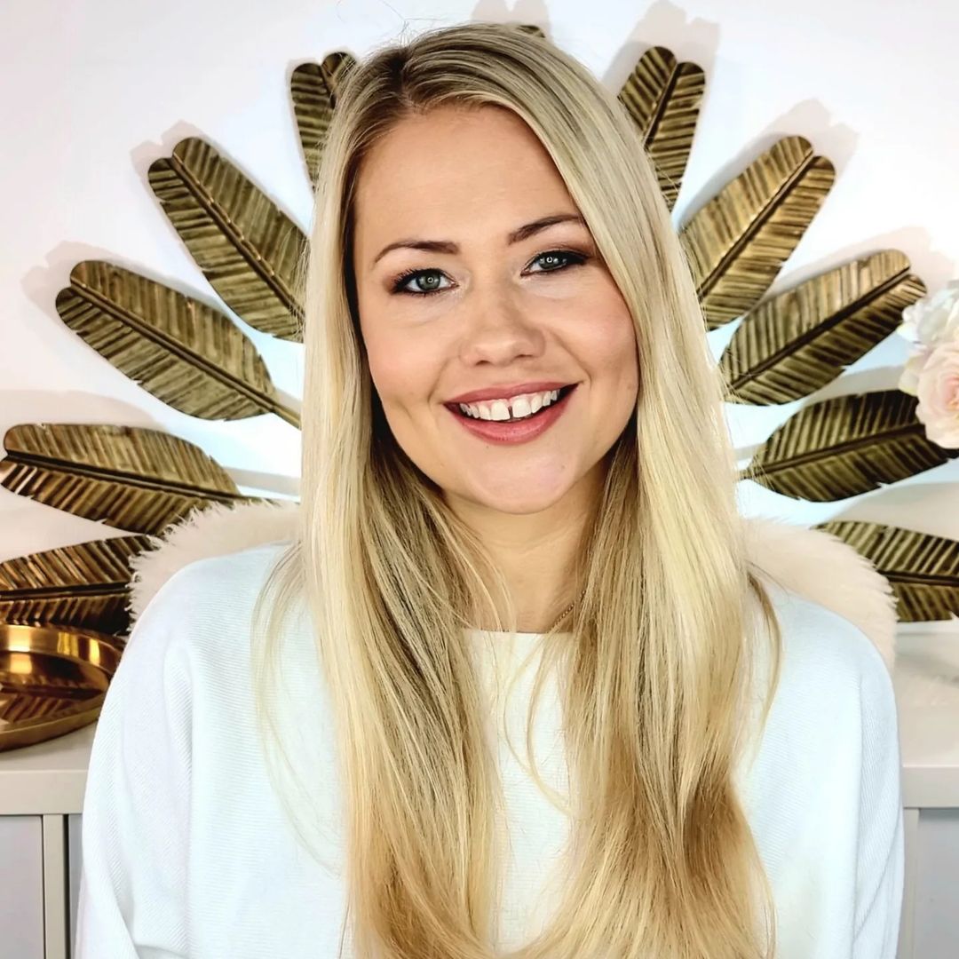Eine Frau mit langen blonden Haaren lächelt vor einer dekorativen Wand mit goldenen Blättern. Sie trägt ein helles Oberteil.