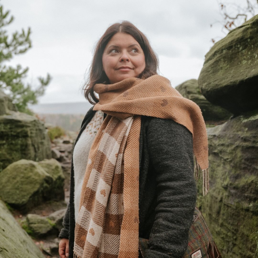 Eine Frau mit braunem Schal und dunklem Mantel steht in einer felsigen Landschaft, umgeben von Bäumen.