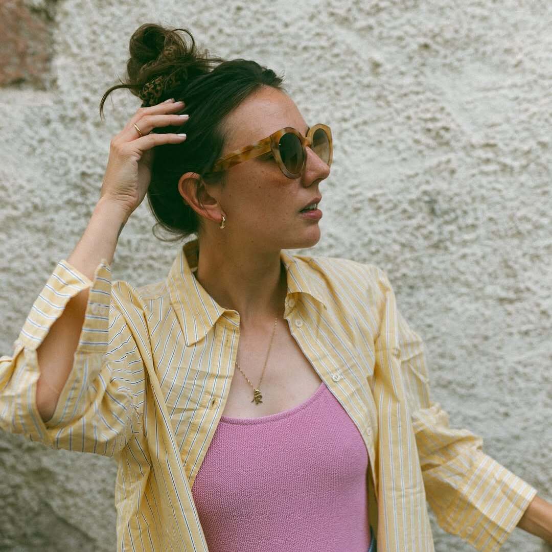 Frau mit Sonnenbrille und gelber Streifenbluse greift sich durch die Haare, lässige Pose vor heller Wand.