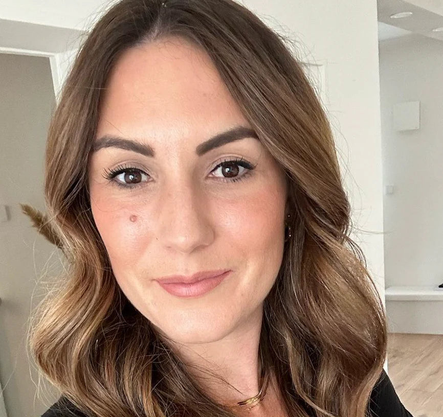 Frau mit schulterlangen, braunen Haaren und sanftem Makeup, vor neutralem Hintergrund.