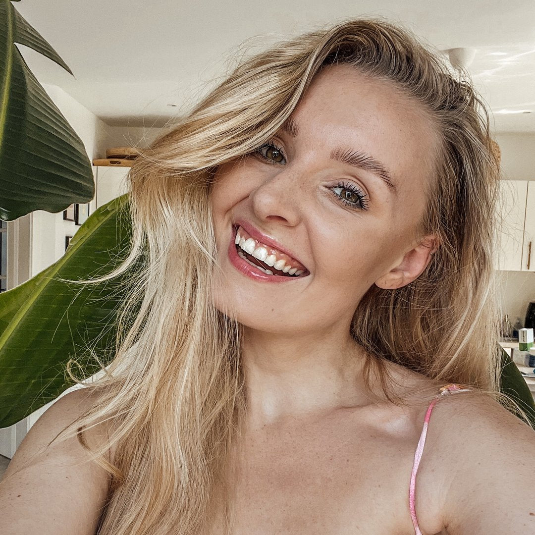Frau mit langen, blonden Haaren lächelt vor großen Pflanzen im Hintergrund. Natürliches Licht und freundliche Atmosphäre.