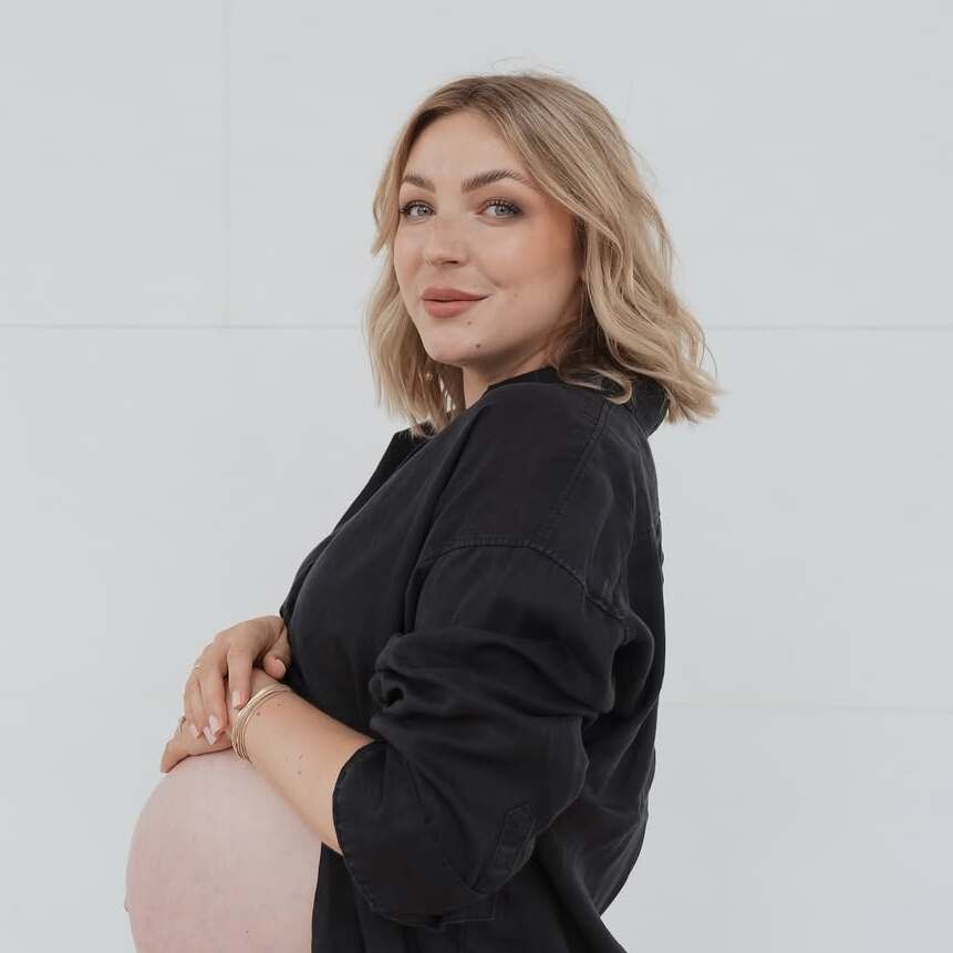 Schwangere Frau in schwarzem Oberteil, die sanft ihren Bauch umarmt, vor neutralem Hintergrund.