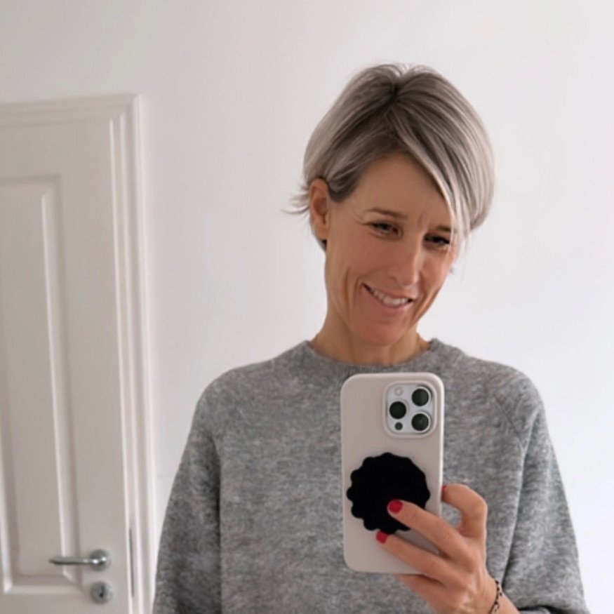 Eine Frau mit kurzen, grauen Haaren lächelt in die Kamera, während sie ein Smartphone in der Hand hält. Sie trägt einen