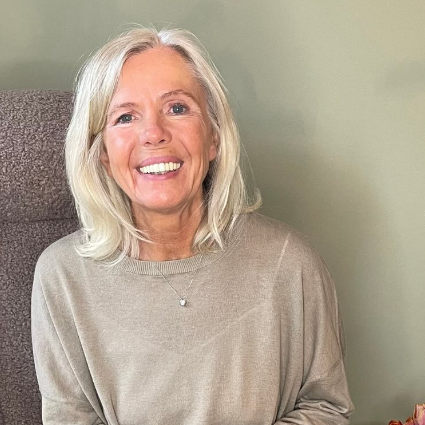 Frau mit blonden, langen Haaren sitzt lächelnd vor einem grauen Hintergrund. Sie trägt ein beige Oberteil und eine Kette.
