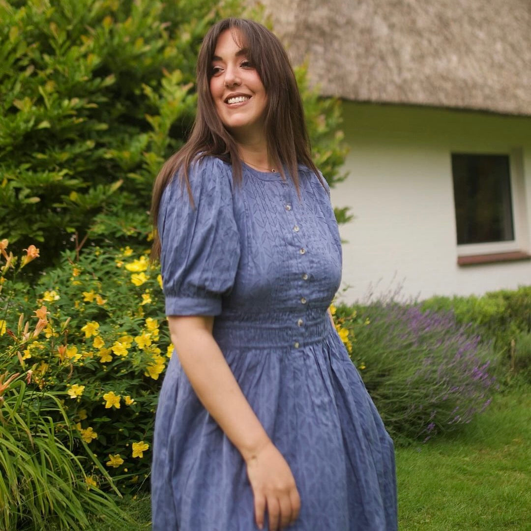 Eine Frau in einem blauen Kleid mit Knöpfen, lächelt in einer blühenden Gartenlandschaft.