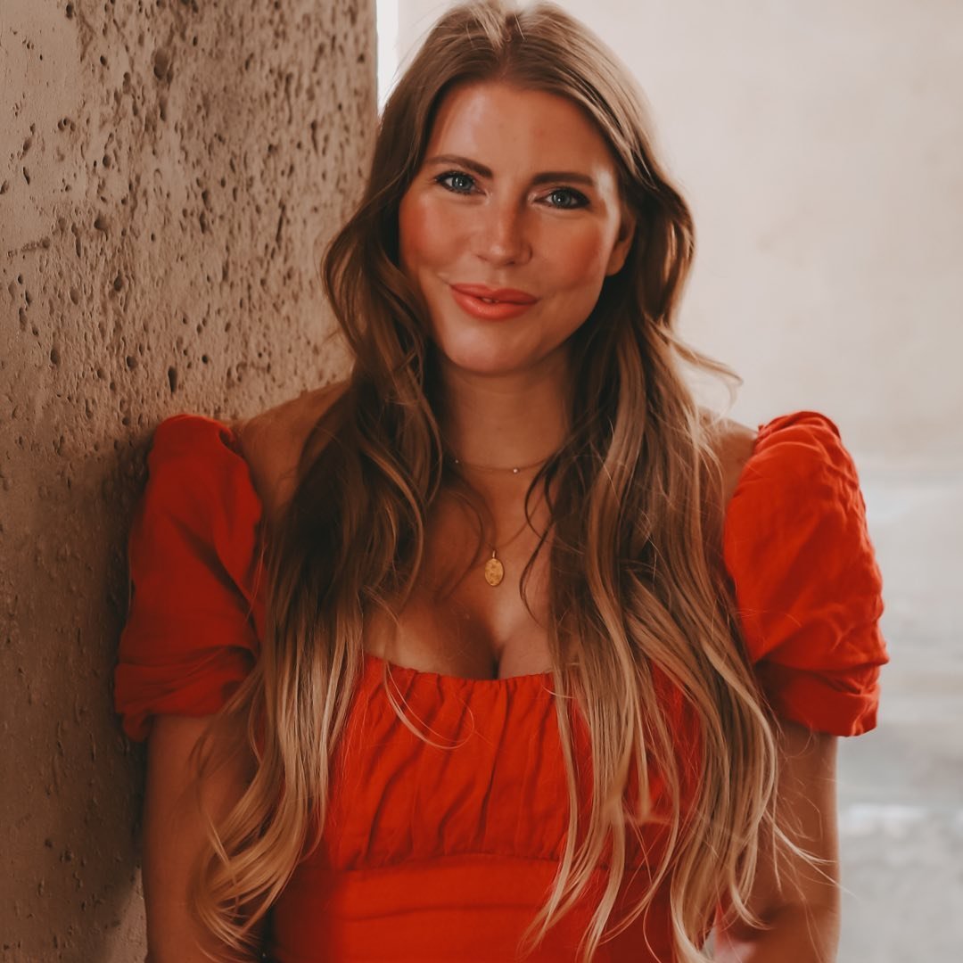 Eine Frau mit langen, lockigen Haaren trägt ein rotes Kleid mit Puffärmeln. Der Hintergrund zeigt eine elegante,