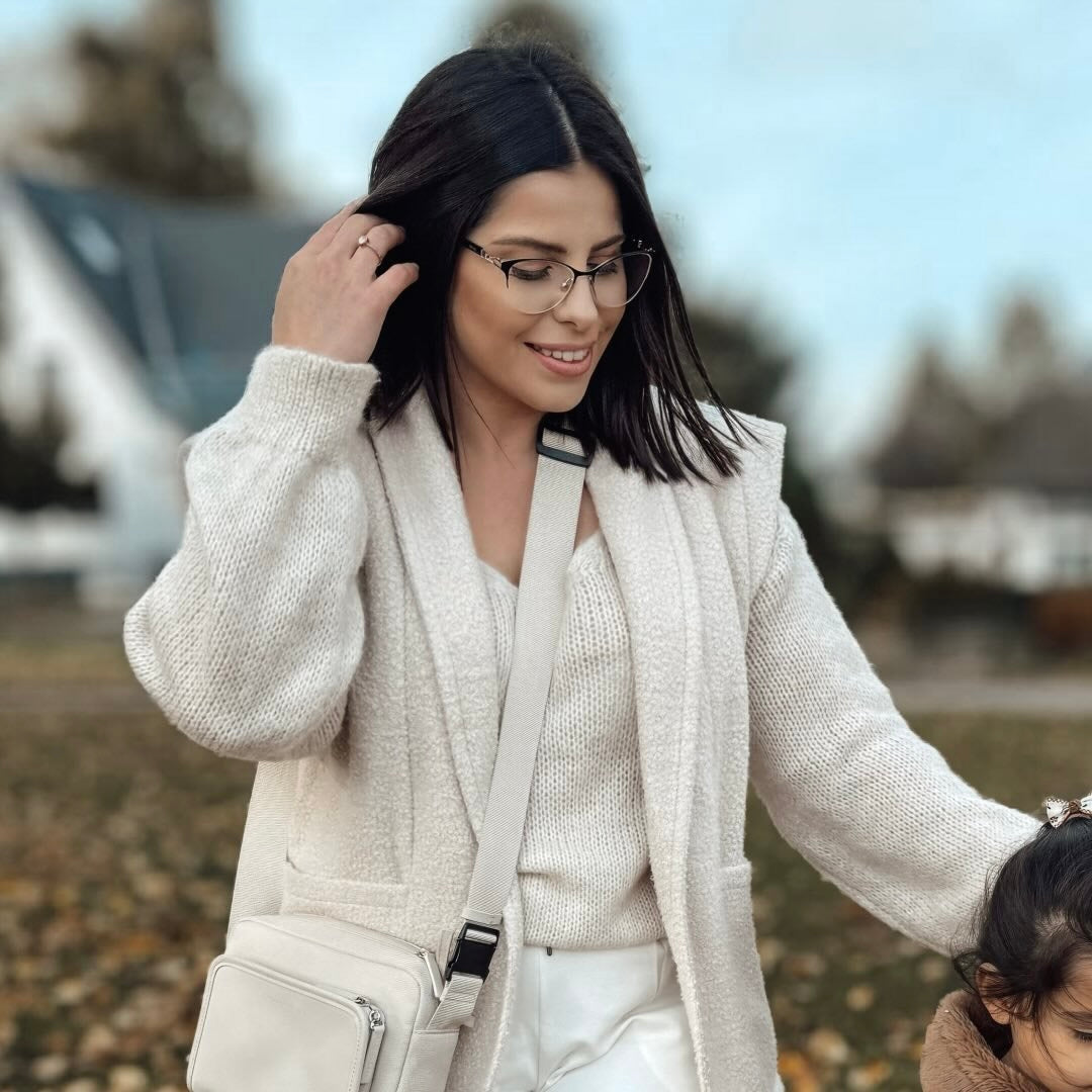 Eine Frau mit Brille in einem gemütlichen, hellen Outfit trägt eine Schultertasche und hält die Hand eines kleinen Mädchens.