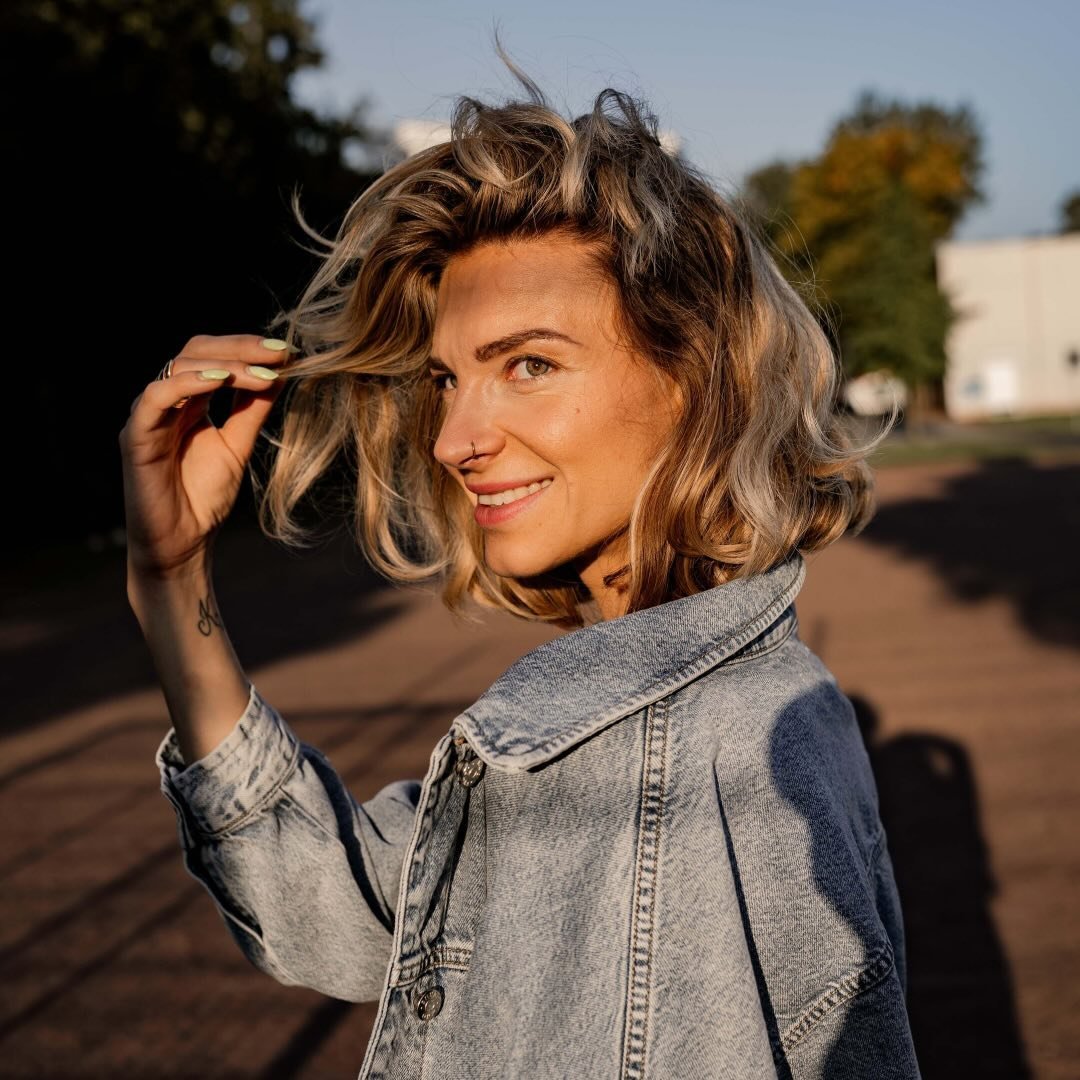 Junge Frau mit lockigem, blondem Haar lächelt in die Kamera, trägt eine Jeansjacke und befindet sich im Freien.