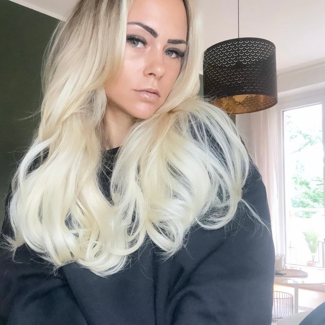 Frau mit langen, blonden Haaren in einem dunklen Pullover steht vor einem Fenster. Im Hintergrund ist eine Lampe sichtbar.