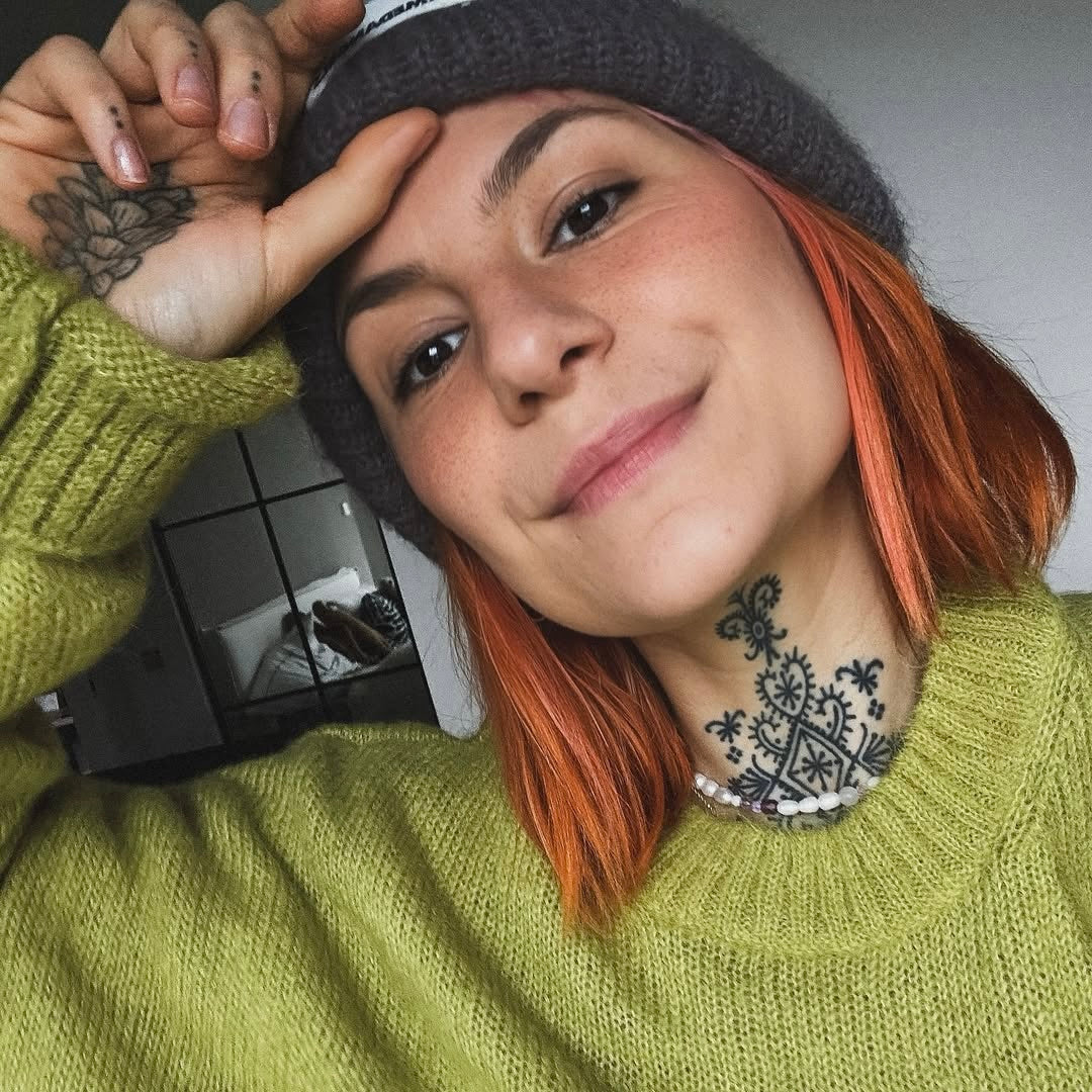 Junge Frau mit orangenem Haar und Strickmütze, lächelt lässig in einem grünen Pullover. Hintergrund unscharf.