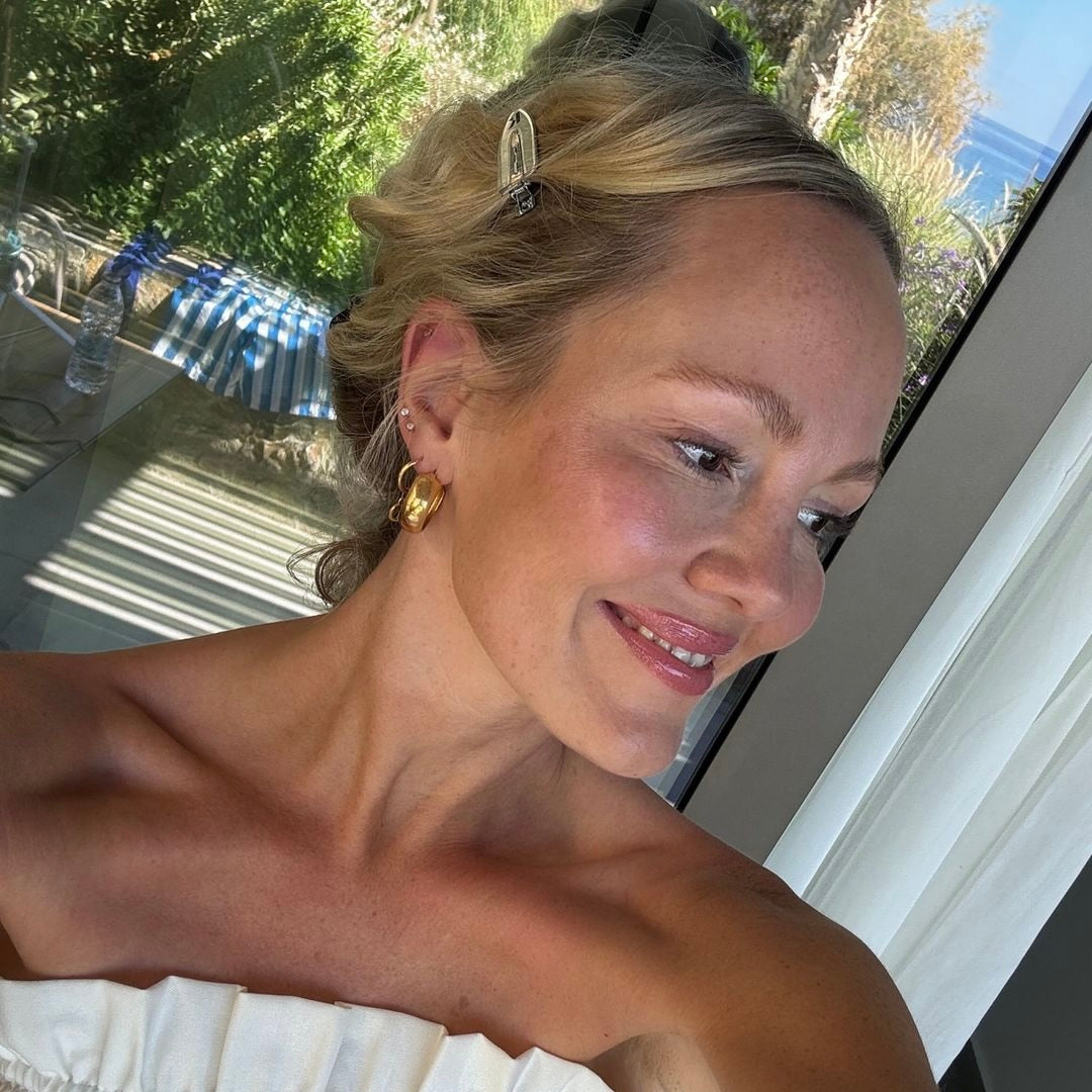 Eine Frau mit hellblonden Haaren, die in einem leichten, trichterförmigen Oberteil posiert. Sie trägt goldene Ohrringe und