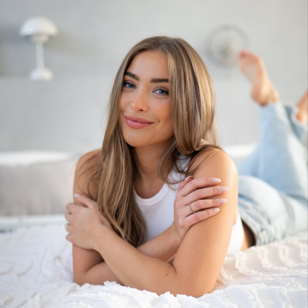 Lächelnde Frau mit langen, glatten Haaren in weißer Kleidung, auf einem Bett liegend. Warme, entspannte Atmosphäre.
