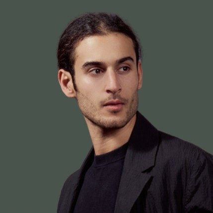 Porträt eines jungen Mannes mit langen, glatten Haaren, der in einem schwarzen Outfit vor einem grauen Hintergrund steht.