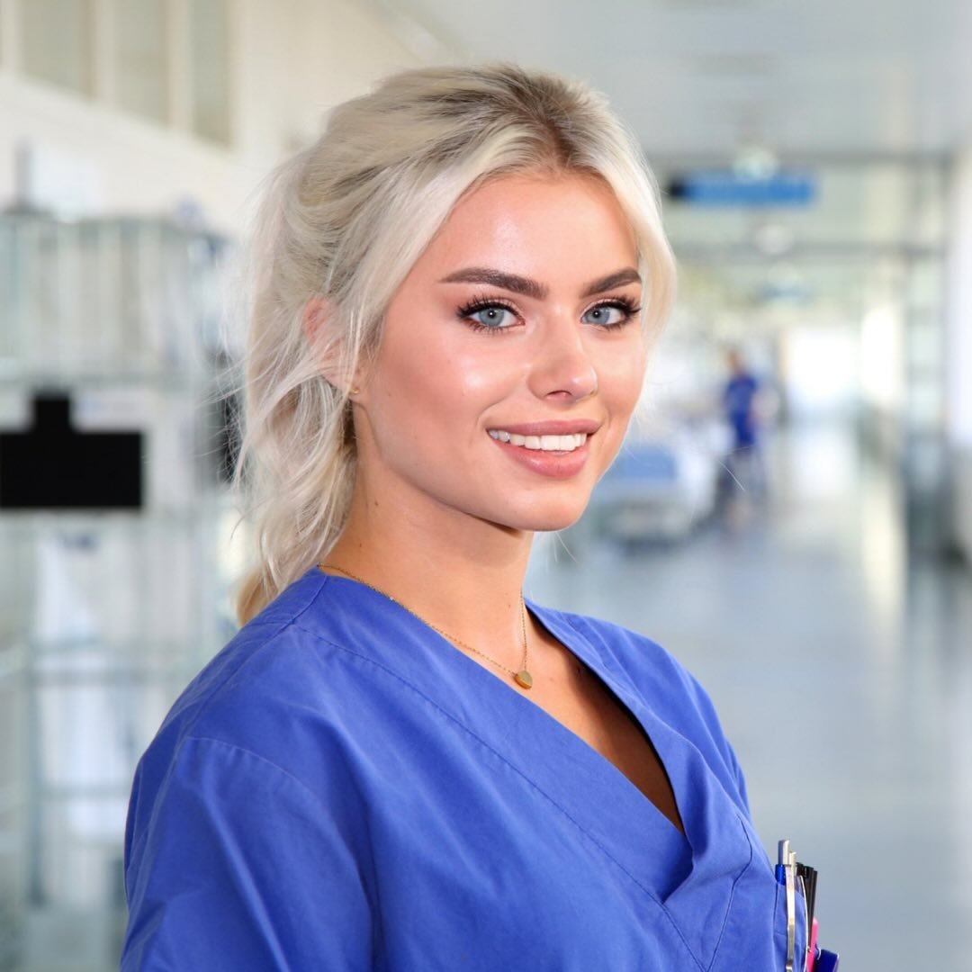 Eine junge Frau mit blonden Haaren und blauen Augen trägt einen blauen OP-Anzug. Sie lächelt freundlich in einer