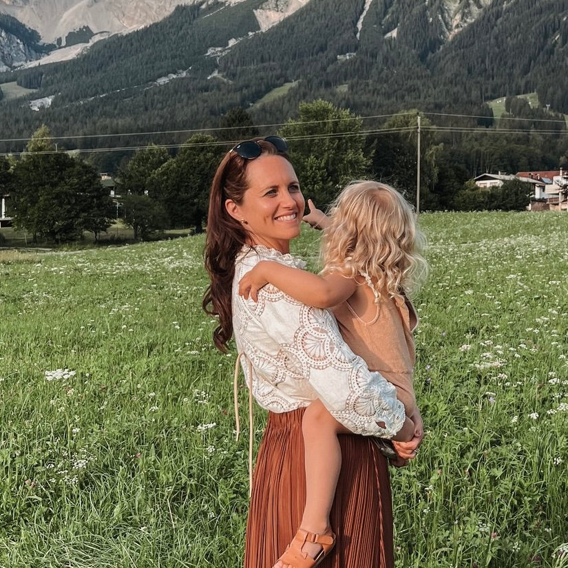 Eine Frau in einem weißen Oberteil hält ein kleines Kind auf dem Arm, umgeben von einer grünen Wiese mit Bergen im