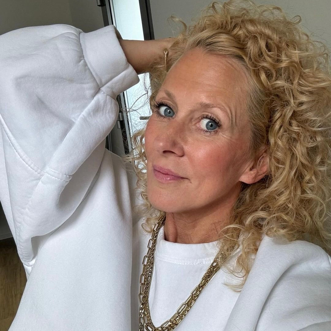 Frau mit lockigem Haar, trägt einen weißen Pullover und eine Kette, lächelt in die Kamera.