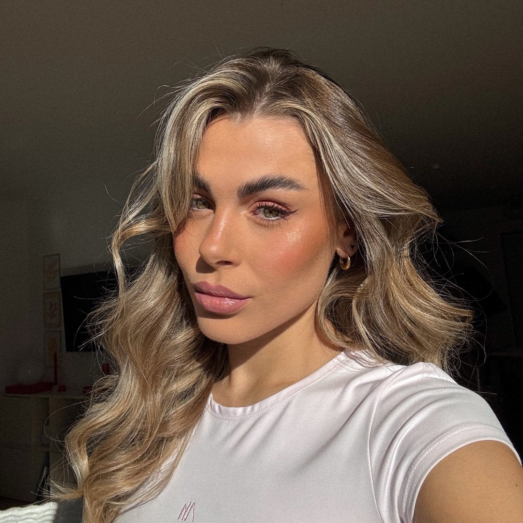 Porträt einer Frau mit gewelltem, blondem Haar und natürlichem Make-up, in einem hellen Raum. Sie trägt ein schlichtes,