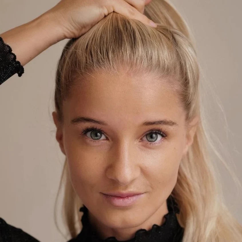 Frau mit langen, blonden Haaren, die ihre Haare nach hinten zieht. Sie trägt ein schwarzes Oberteil und schaut direkt in die