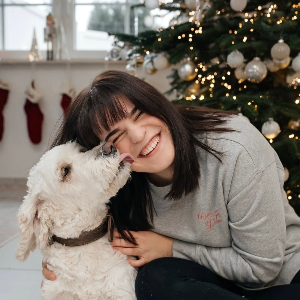 Junge Frau mit braunen Haaren, die lächelt und mit einem weißen Hund spielt, vor einem geschmückten Weihnachtsbaum.