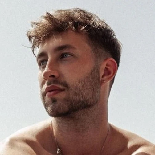 Ein junger Mann mit kurzer, zerzauster Frisur, oberkörperfrei, schaut nachdenklich in die Ferne. Heller Hintergrund.