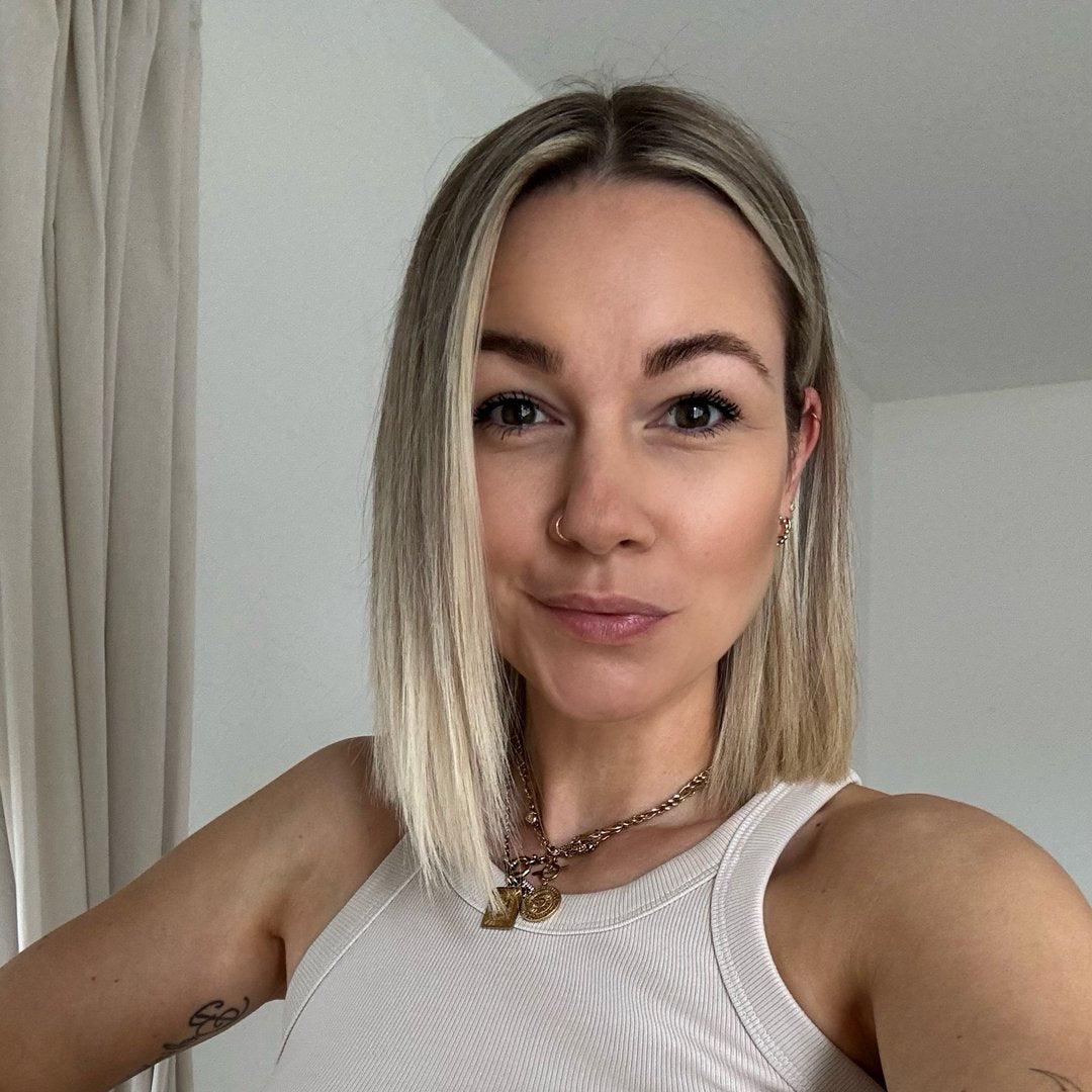 Eine Frau mit blonden, schulterlangen Haaren und einem schlichten Tanktop lächelt in die Kamera. Ihr Look ist lässig und