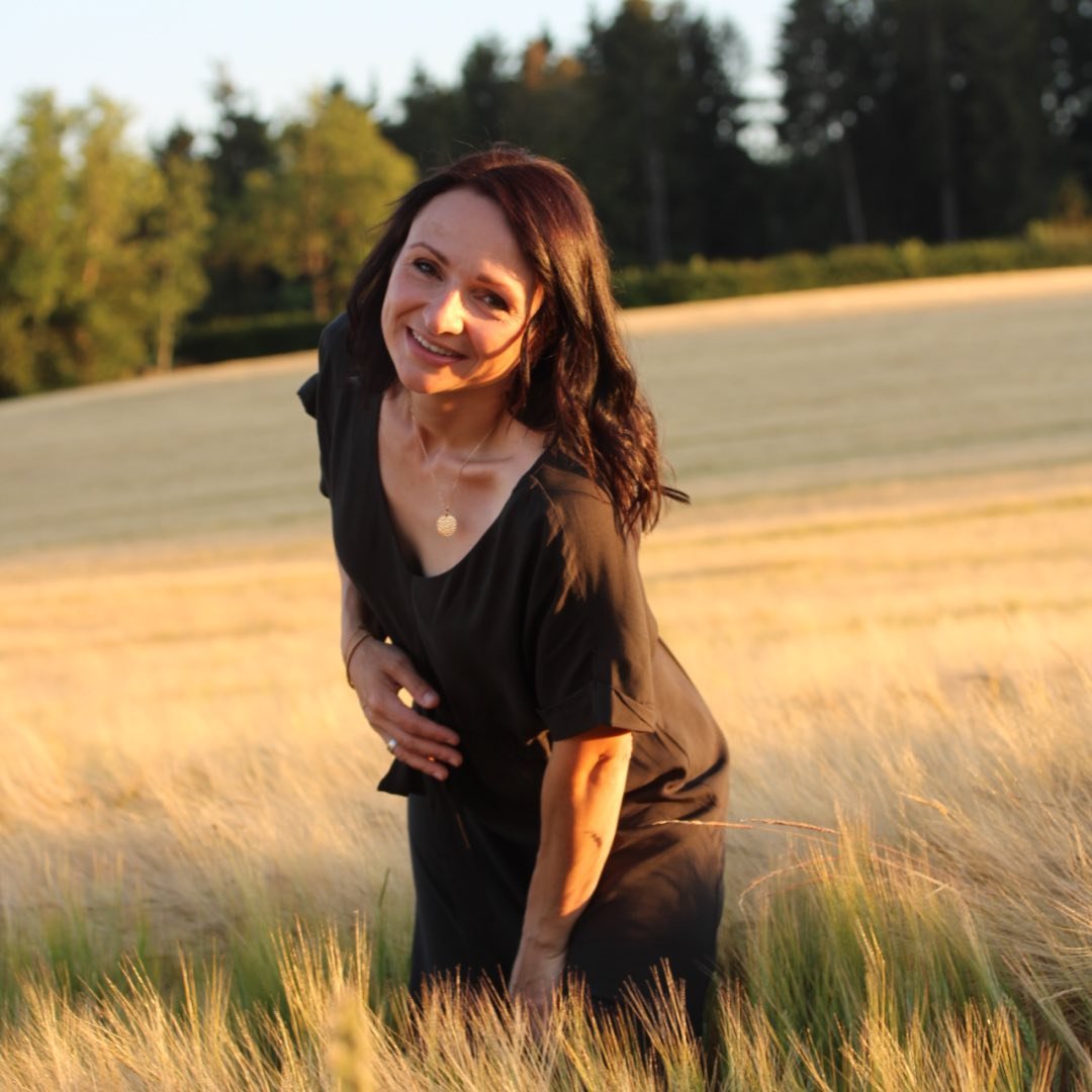 Eine Frau in einem Feld mit hohem Gras, lächelt entspannt in die Kamera. Sie trägt ein schlichtes, dunkles Kleid.
