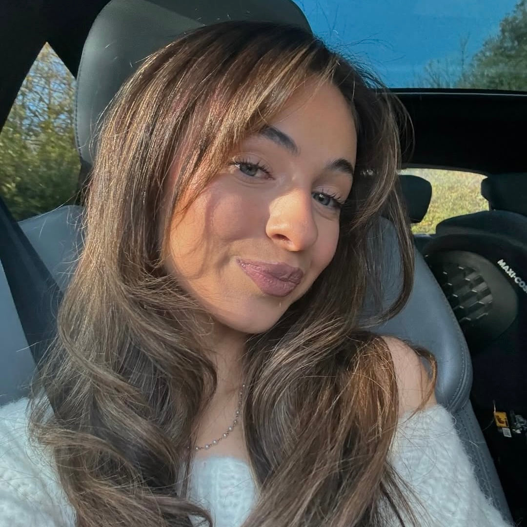 Eine Frau mit langen, braunen Haaren und einem sanften Lächeln, sitzt im Auto mit einem hellen Pulli.