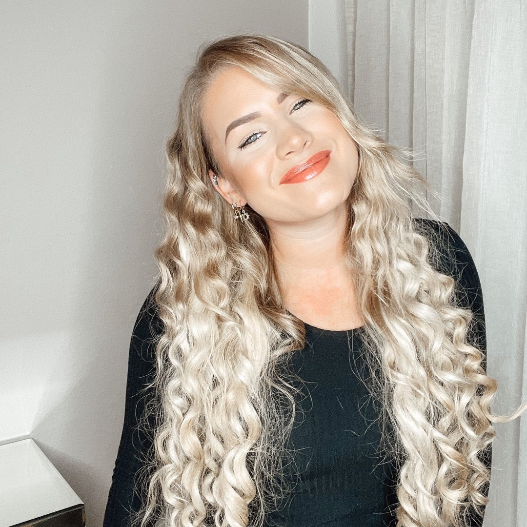 Lächelnde Frau mit langen, lockigen blonden Haaren in Schwarz, fröhlich in einem hellen Raum.