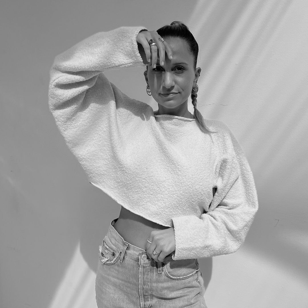 Frau in einem hellen, lockeren, cropped Pullover zeigt eine Pose. Der Hintergrund ist hell mit Schatten. Der Stil ist modern