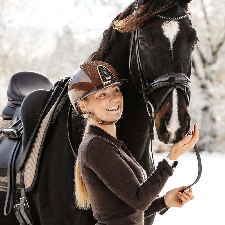 Eine Frau mit einem braunen Reithelm steht neben einem schwarzen Pferd und lächelt. Die Winterlandschaft im Hintergrund ist