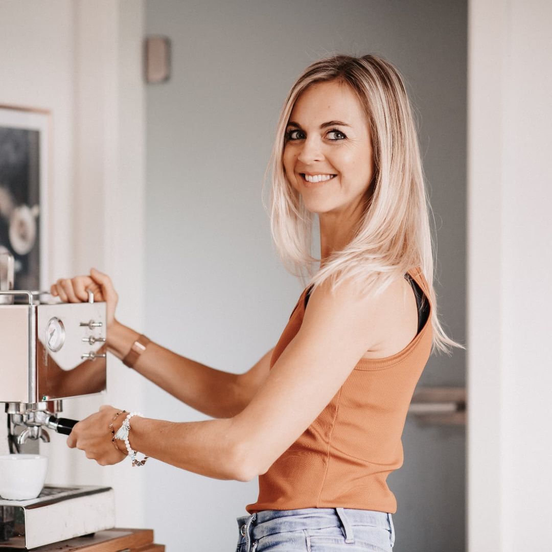Eine Frau mit blonden Haaren steht an einer Espressomaschine und bereitet Kaffee zu. Sie trägt ein oranges Top und wirkt