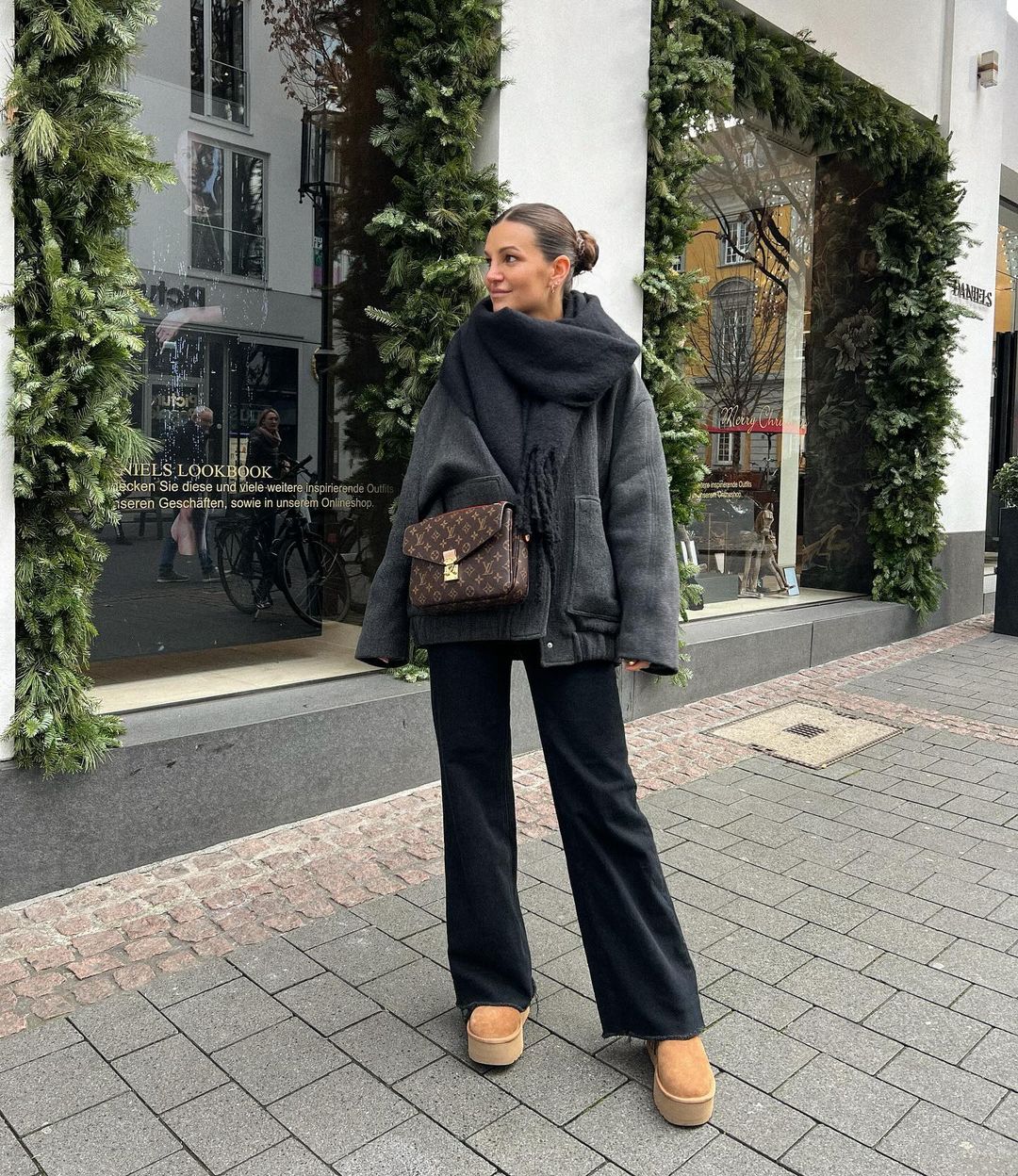 Eine Frau trägt einen grauen Mantel, schwarze Hose und Boots, steht vor einem Geschäft, umgeben von Weihnachtsdekoration.