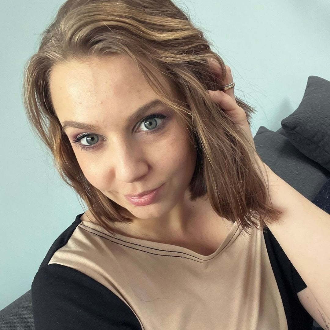 Eine Frau mit schulterlangen, lockigen Haaren sitzt auf einer Couch und schaut in die Kamera. Sie trägt ein beiges Oberteil