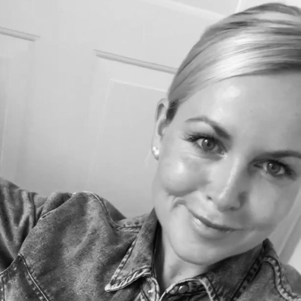 Lächelnde Frau mit blonder Haarfrisur in Jeansjacke vor heller Wand.