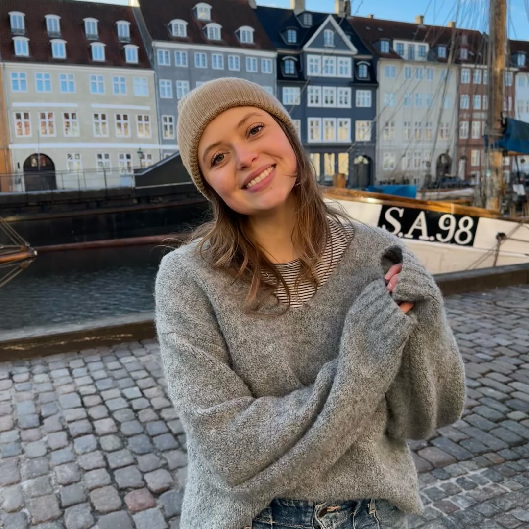 Eine Frau trägt einen beigen Hut und einen grauen, lockeren Pullover. Im Hintergrund sind historische Gebäude und ein Boot