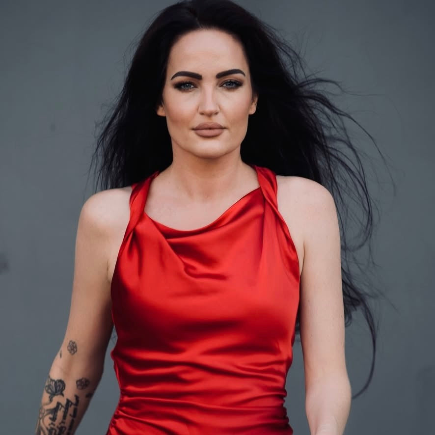 Frau mit langen schwarzen Haaren trägt ein rotes satinähnliches Kleid vor grauem Hintergrund.