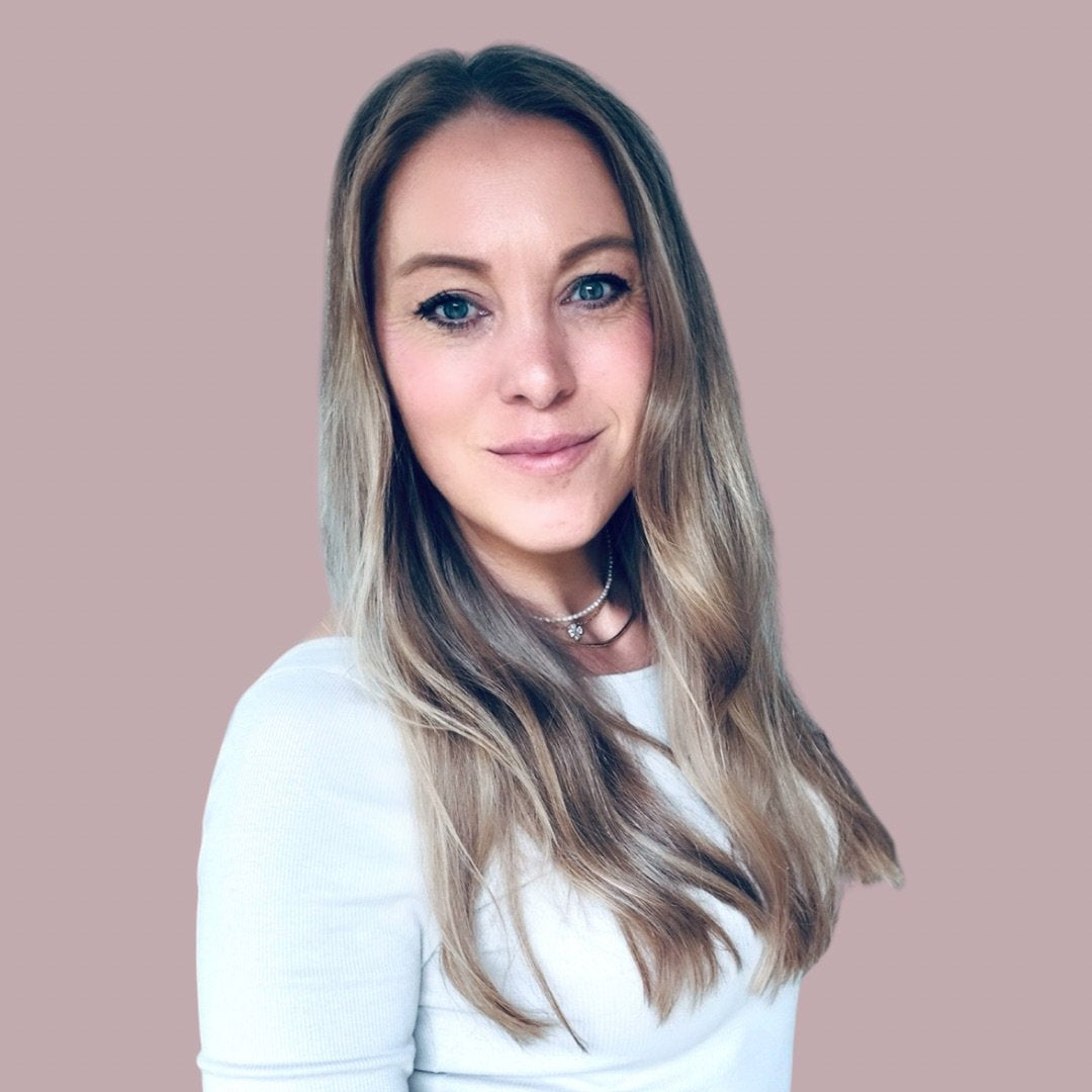 Eine Frau mit langen, welligen Haaren und einem dezenten Choker, lächelt vor einem sanften, einfarbigen Hintergrund.