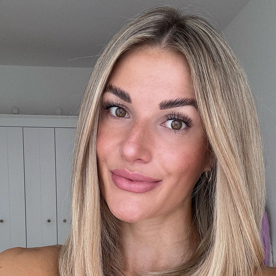 Portrait einer Frau mit blonden, langen Haaren und sanftem Lächeln vor einem hellen Hintergrund.