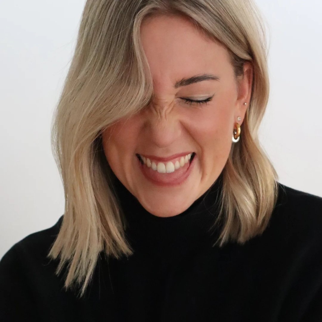 Frau mit platinblonden Haaren und dunklem Pullover, lächelt fröhlich und strahlt Wärme aus.
