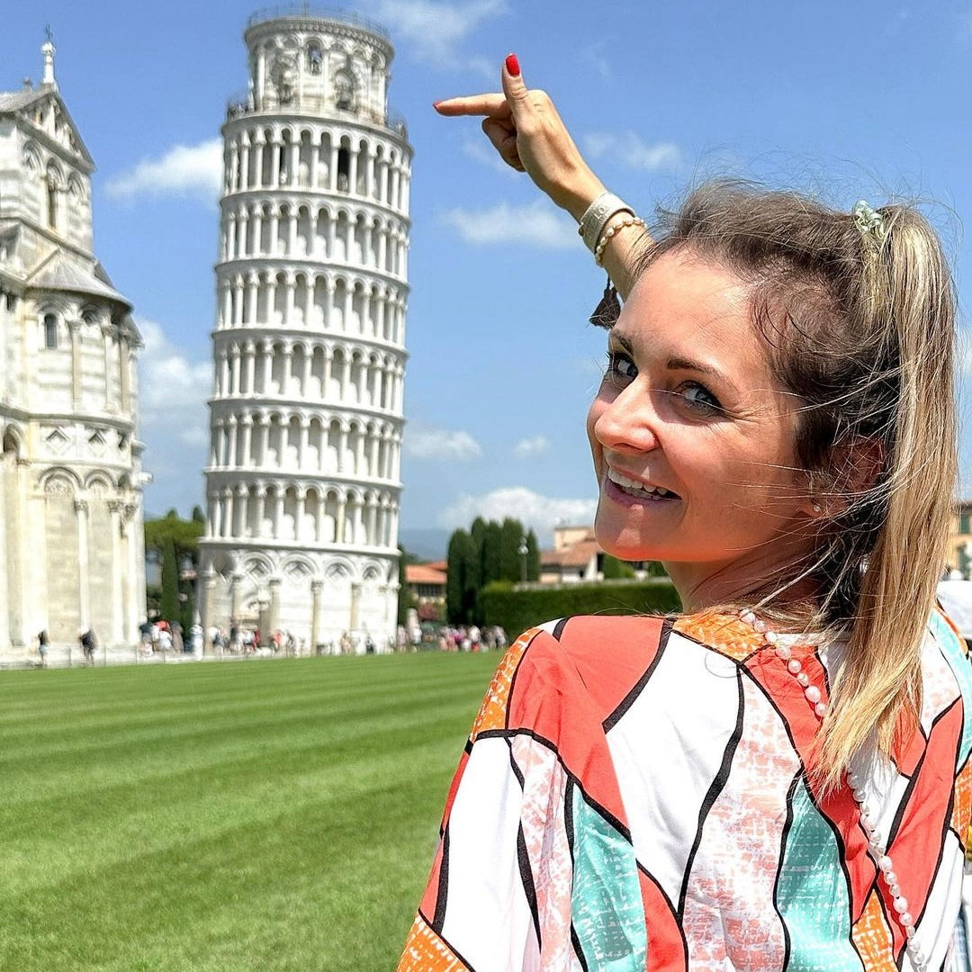 Frau in bunter Bluse zeigt auf den Schiefen Turm von Pisa, umgeben von grünen Wiesen und blauem Himmel.