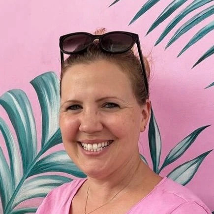 Eine Frau mit Sonnenbrille vor einer pinken Wand mit Blattmotiven. Sie lächelt freundlich.