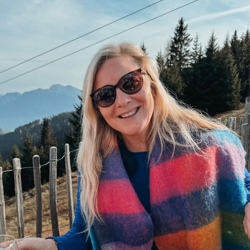 Eine lächelnde Frau mit Sonnenbrille trägt einen bunten Schal. Im Hintergrund sind Berge und Bäume sichtbar.