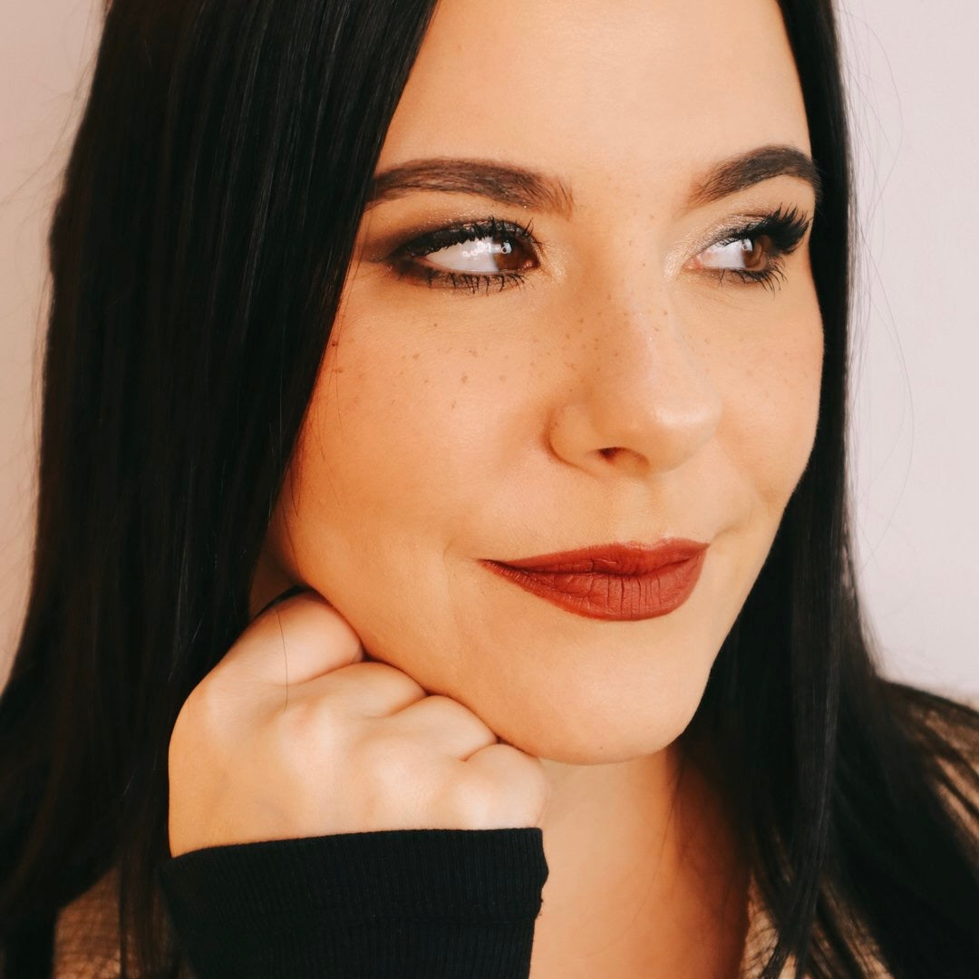 Porträt einer Frau mit dunklen Haaren und rotem Lippenstift. Sie lächelt leicht und stützt ihr Kinn mit der Hand ab.