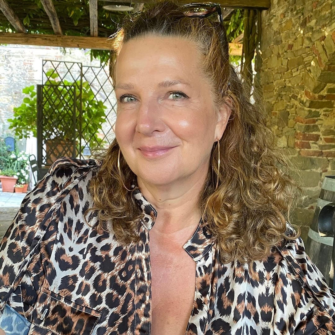 Frau mit lockigem Haar trägt ein leopardenmusteriertes Oberteil. Natürliche Umgebung mit Pflanzen im Hintergrund.