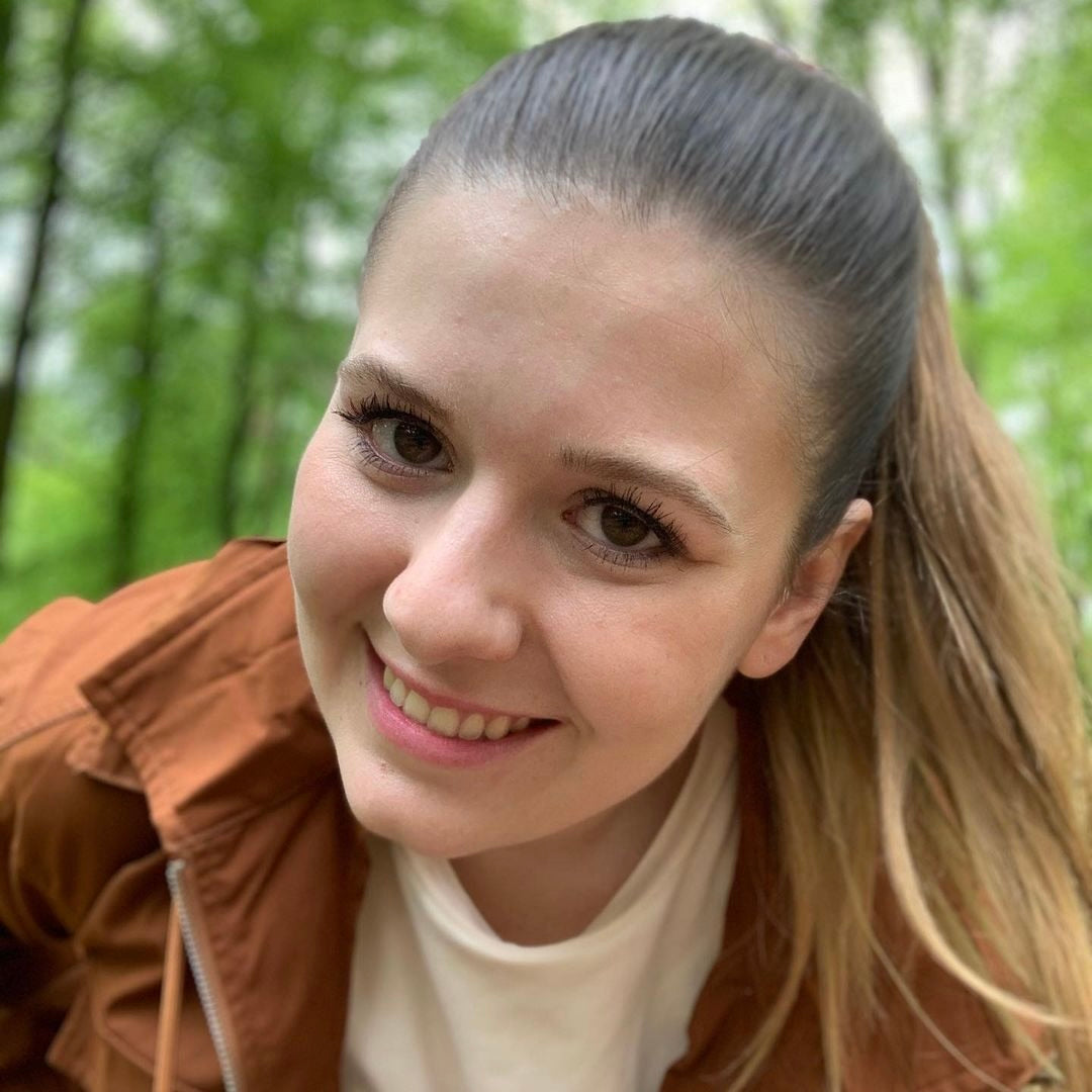 Junge Frau mit langen, glatten Haaren und freundlichem Lächeln in der Natur, grüne Bäume im Hintergrund.