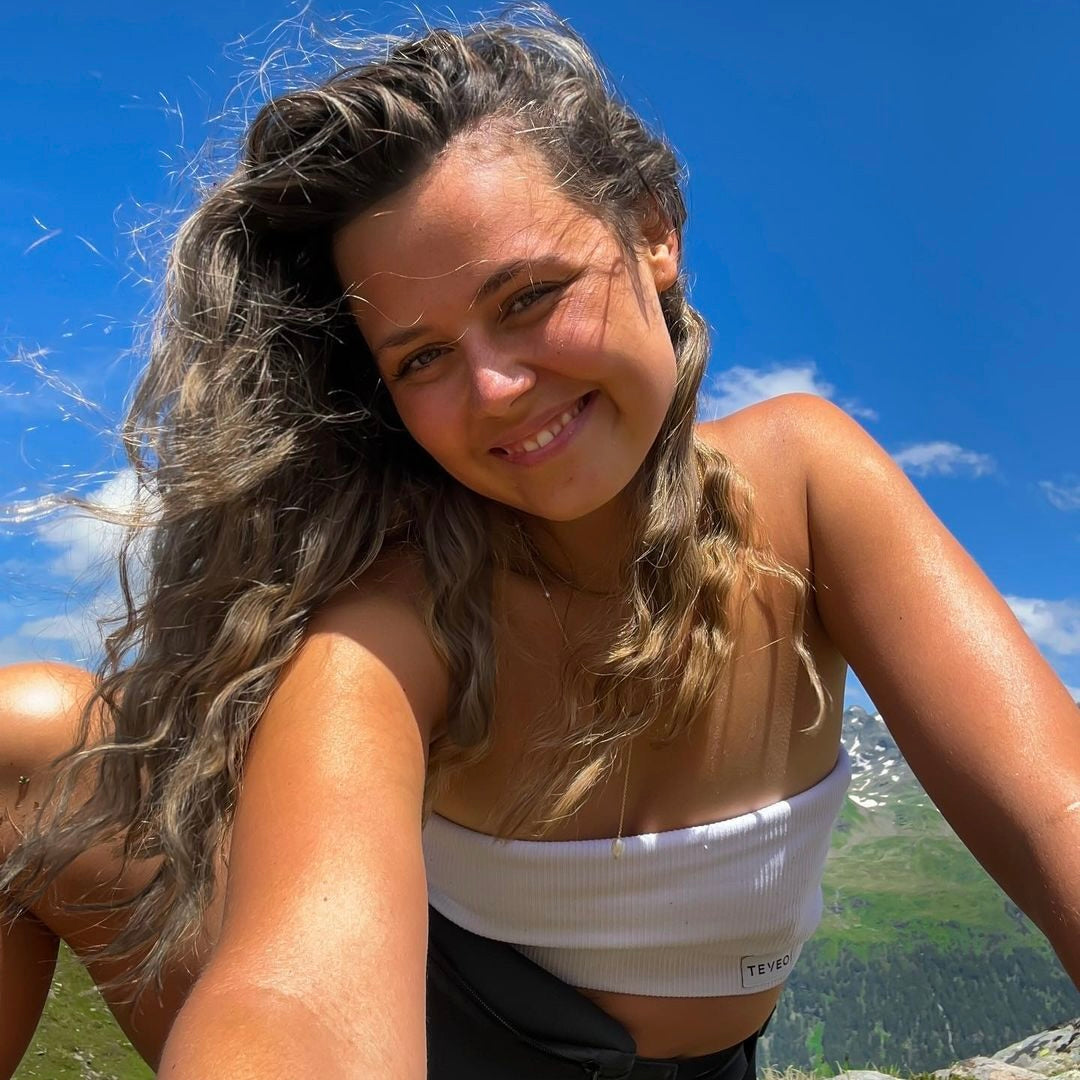 Frau mit lockigem, brünettem Haar sitzt in der Natur bei Sonnenschein, lächelnd und entspannt.