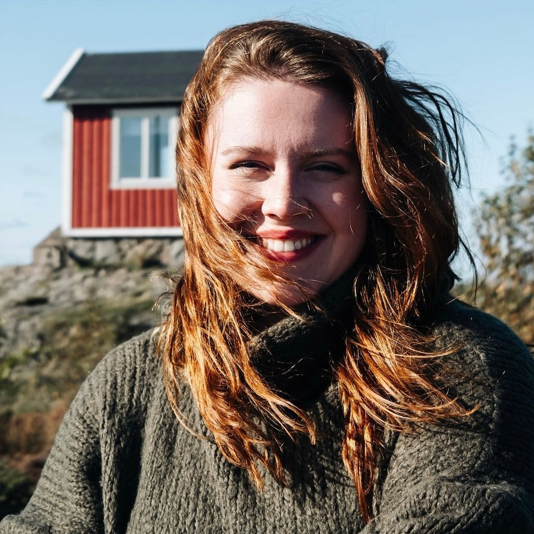 Frau mit langen, lockigen roten Haaren trägt einen dunklen Pullover. Hinter ihr steht ein rotes Holzhaus vor blauer Himmel.