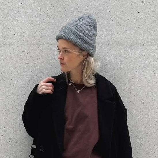 Frau mit grauer Mütze und schwarzer Jacke vor einer hellen Wand. Ihr Blick ist nachdenklich, die Haare sind leicht gewellt.