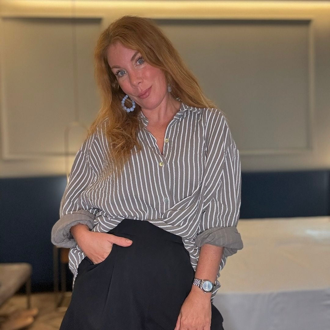 Frau mit langen roten Haaren in einem gestreiften Hemd und schwarzer Hose, lächelt in einem modernen Raum.