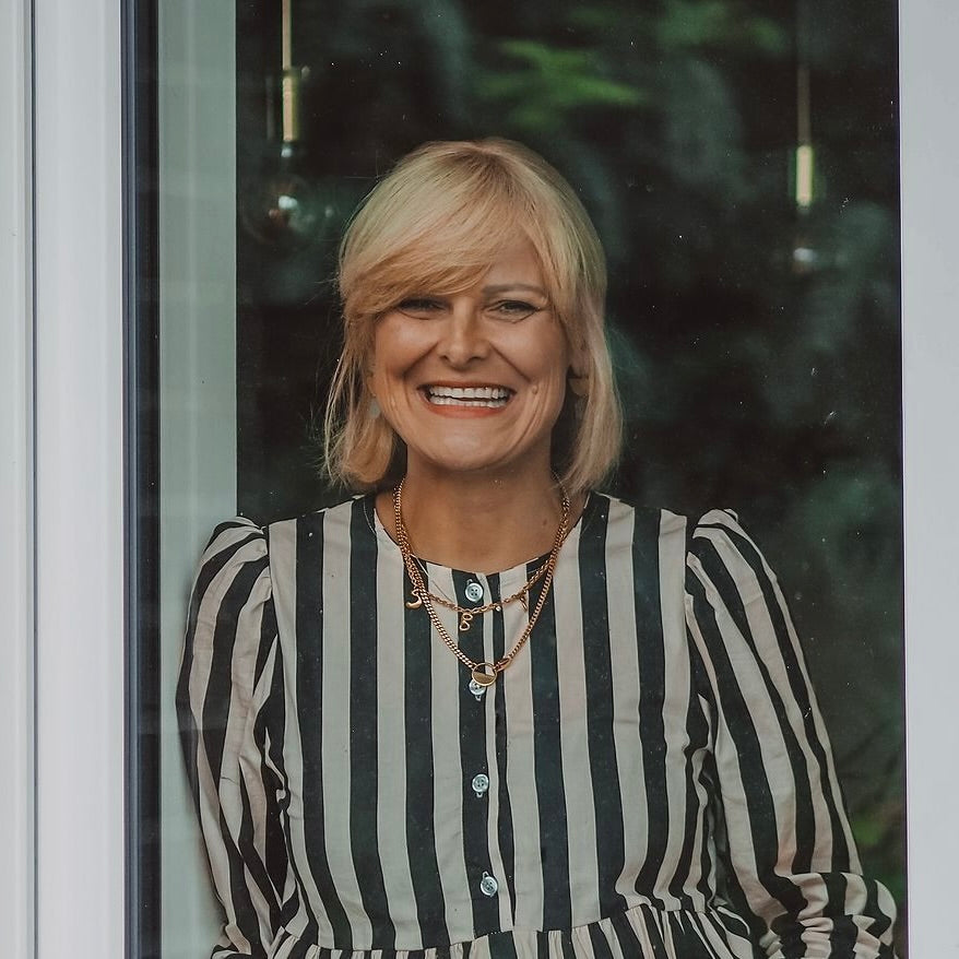 Frau mit blonden, schulterlangen Haaren, lächelnd hinter einem Fenster. Sie trägt ein gestreiftes, langärmeliges Kleid.